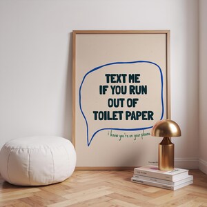 May include: A framed print with a blue speech bubble that says "TEXT ME IF YOU RUN OUT OF TOILET PAPER" and the text "i know you're on your phone" underneath. The print is on a beige background and is framed in a light wood frame.