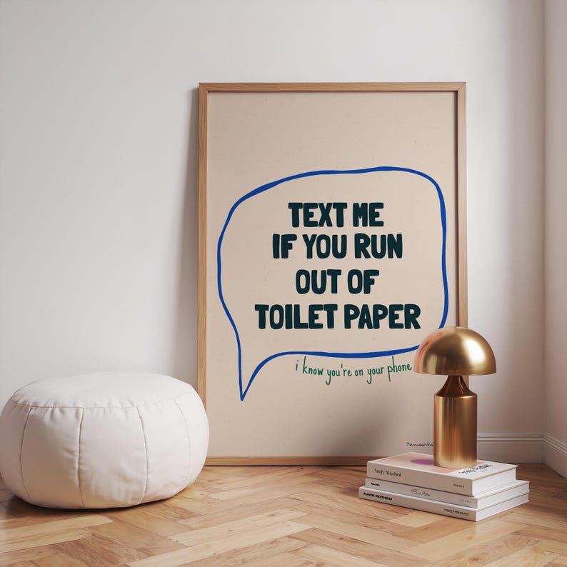 May include: A framed print with a blue speech bubble that says "TEXT ME IF YOU RUN OUT OF TOILET PAPER" and the text "i know you're on your phone" underneath. The print is on a beige background and is framed in a light wood frame.