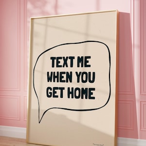 May include: A beige poster with a black speech bubble that says "Text me when you get home". The poster is framed in gold and is hanging on a pink wall.