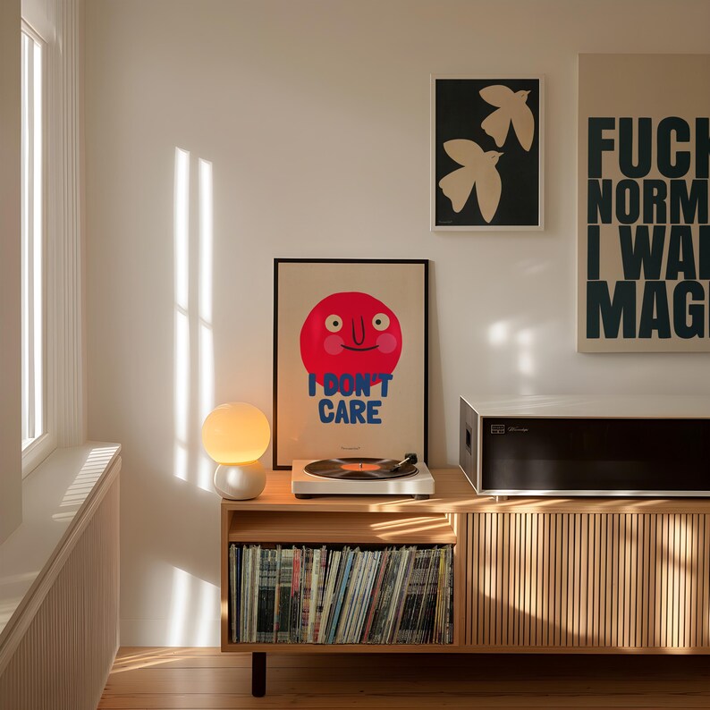 May include: A wooden record player stand with a record player, a lamp, and a collection of vinyl records. The stand has a light wood finish and a slatted front. There are three posters on the wall behind the stand. One poster features a red smiling face with the text "I don't care". Another poster features a black and white illustration of two birds. The third poster features the text "F*ck Norm I Want Magic".
