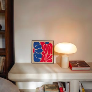 May include: A white table lamp with a mushroom-shaped shade sits on a white shelf next to a framed print of a red and blue abstract floral design.