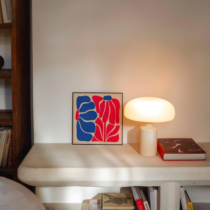 May include: A white table lamp with a mushroom-shaped shade sits on a white shelf next to a framed print of a red and blue abstract floral design.