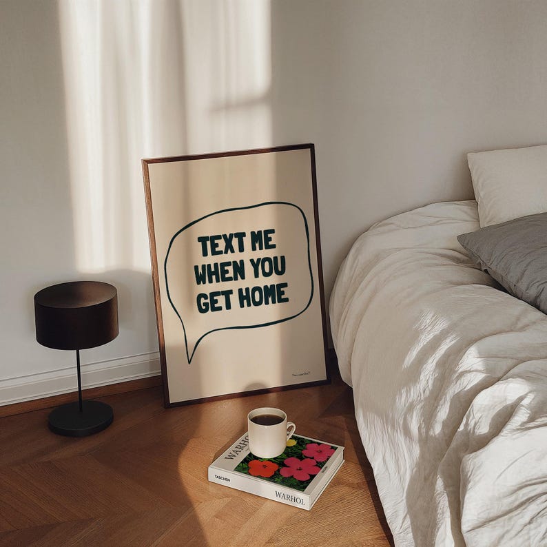 May include: A framed print with a white background and black text that reads "Text me when you get home" in a speech bubble. The print is sitting on a wooden floor next to a white bed with a white blanket and a book with a floral design.
