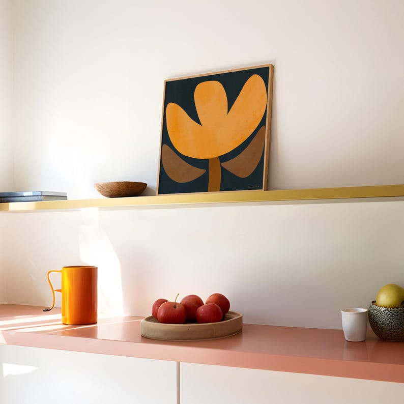 May include: A square framed print of an abstract flower with orange, brown, and dark blue colors on a white wall above a shelf with a wooden bowl and books.