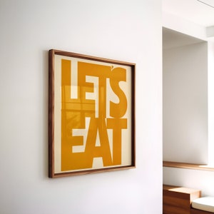 以下が含まれることがあります： 太字のオレンジ色の文字で「LET'S EAT」と書かれた額入りのアートプリント。 アートワークはクリーム色の背景に設定され、濃い茶色の木製フレームに収められています。 プリントは白い壁に掛けられています。