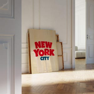 May include: A white canvas with the words "NEW YORK CITY" in red and blue letters. The canvas is leaning against a white wall in a room with wooden floors.