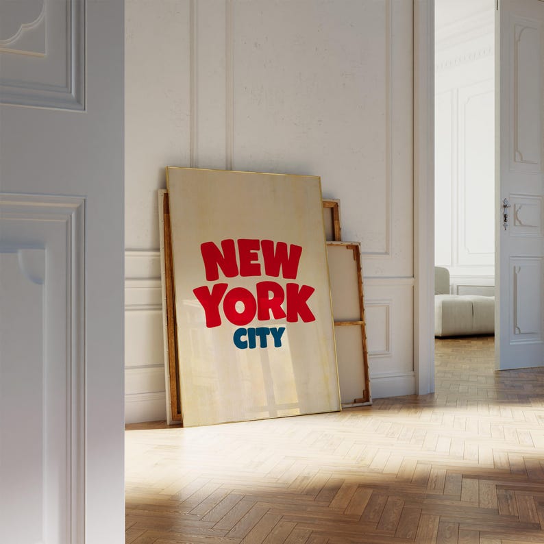 May include: A white canvas with the words "NEW YORK CITY" in red and blue letters. The canvas is leaning against a white wall in a room with wooden floors.