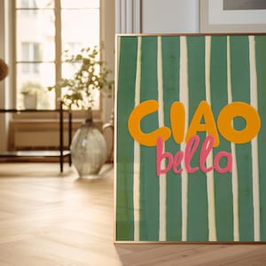 May include: A framed art print with a green and white striped background. The words "CIAO bella" are written in orange and pink. The artwork is displayed on a wooden floor in a room with natural light.