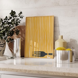 Op de afbeelding: Een ingelijst print met een gele en witte gestreepte achtergrond en een zwart silhouet van een vork met spaghetti erop. De print staat op een wit aanrecht met een zilveren kan en een zilveren kom met gele citroenen.