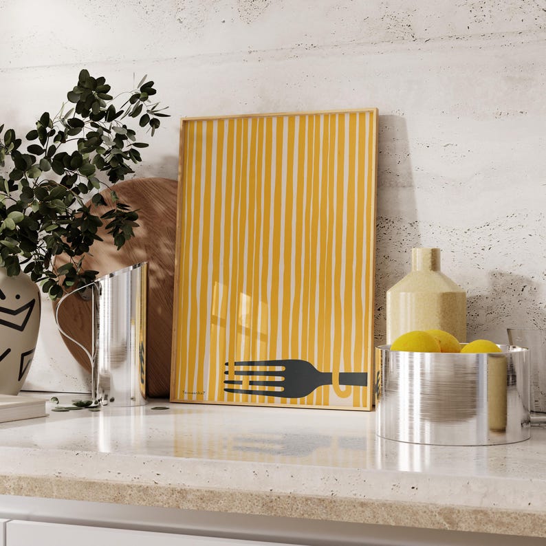 Op de afbeelding: Een ingelijst print met een gele en witte gestreepte achtergrond en een zwart silhouet van een vork met spaghetti erop. De print staat op een wit aanrecht met een zilveren kan en een zilveren kom met gele citroenen.