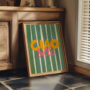 May include: A framed art print with a green and white striped background. The words "CIAO bella" are in orange and pink. The frame is a light brown color. The artwork is leaning against a wooden cabinet.