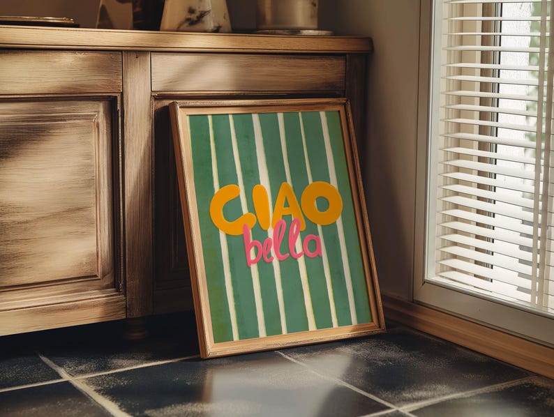 May include: A framed art print with a green and white striped background. The words "CIAO bella" are in orange and pink. The frame is a light brown color. The artwork is leaning against a wooden cabinet.