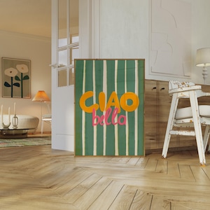 May include: A framed art print featuring the words "CIAO bella" in orange and pink against a green and white striped background. The artwork is displayed in a well-lit room with wooden flooring.