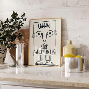 May include: A framed poster with a cartoon illustration of a person holding a sign that says "Stop Overthinking". The person has a simple face with two eyes and a smile. The poster has a light beige background and a wooden frame.