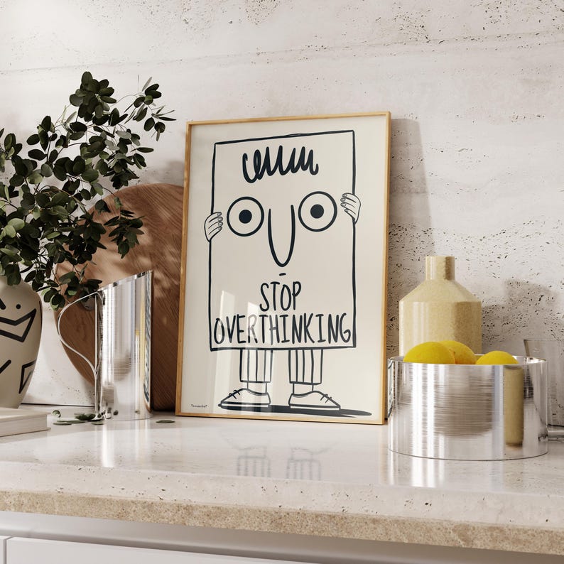 May include: A framed poster with a cartoon illustration of a person holding a sign that says "Stop Overthinking". The person has a simple face with two eyes and a smile. The poster has a light beige background and a wooden frame.