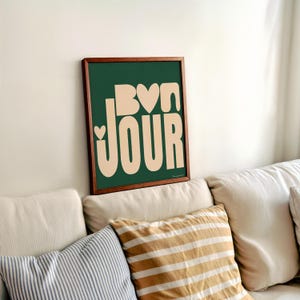May include: A framed print with a green background and the word "BONJOUR" in a peach color with hearts in the letters. The print is hanging on a white wall above a white couch with pillows.