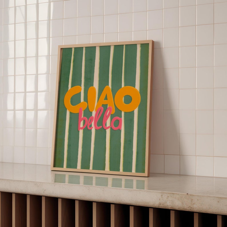 May include: A framed art print with a green and white striped background. The words "CIAO bella" are in orange and pink. The artwork is displayed on a white tiled wall.