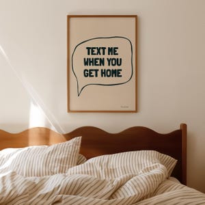 May include: A framed print with a white background and a black speech bubble that says "Text me when you get home". The print is hanging on a white wall above a bed with a wooden headboard and striped bedding.