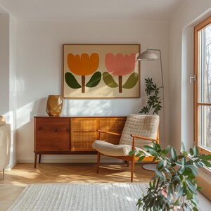 Może przedstawiać: Salon w stylu mid-century modern z drewnianym kredensem, białym fotelem i dużym, oprawionym wydrukiem dw&oacute;ch stylizowanych kwiat&oacute;w w kolorze pomarańczowym, r&oacute;żowym i zielonym. Widoczna jest r&oacute;wnież lampa stojąca z białym kloszem i doniczkowa roślina.