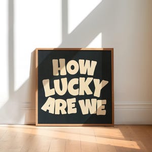 May include: A framed art print with the words "HOW LUCKY ARE WE" in a bold, cream-colored font against a black background. The frame is a warm brown color, and the artwork is displayed on a wooden floor.