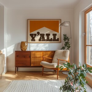 May include: A mid-century modern living room with a wooden cabinet, a white armchair, and a large framed print with the word "Y'ALL" in bold black letters on a yellow and white background.