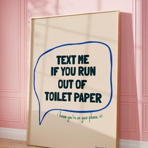May include: A beige poster with a blue speech bubble that says "TEXT ME IF YOU RUN OUT OF TOILET PAPER" in black text. Below the speech bubble, it says "I know you're on your phone =)" in green text. The poster is framed in gold and is hanging on a pink wall.