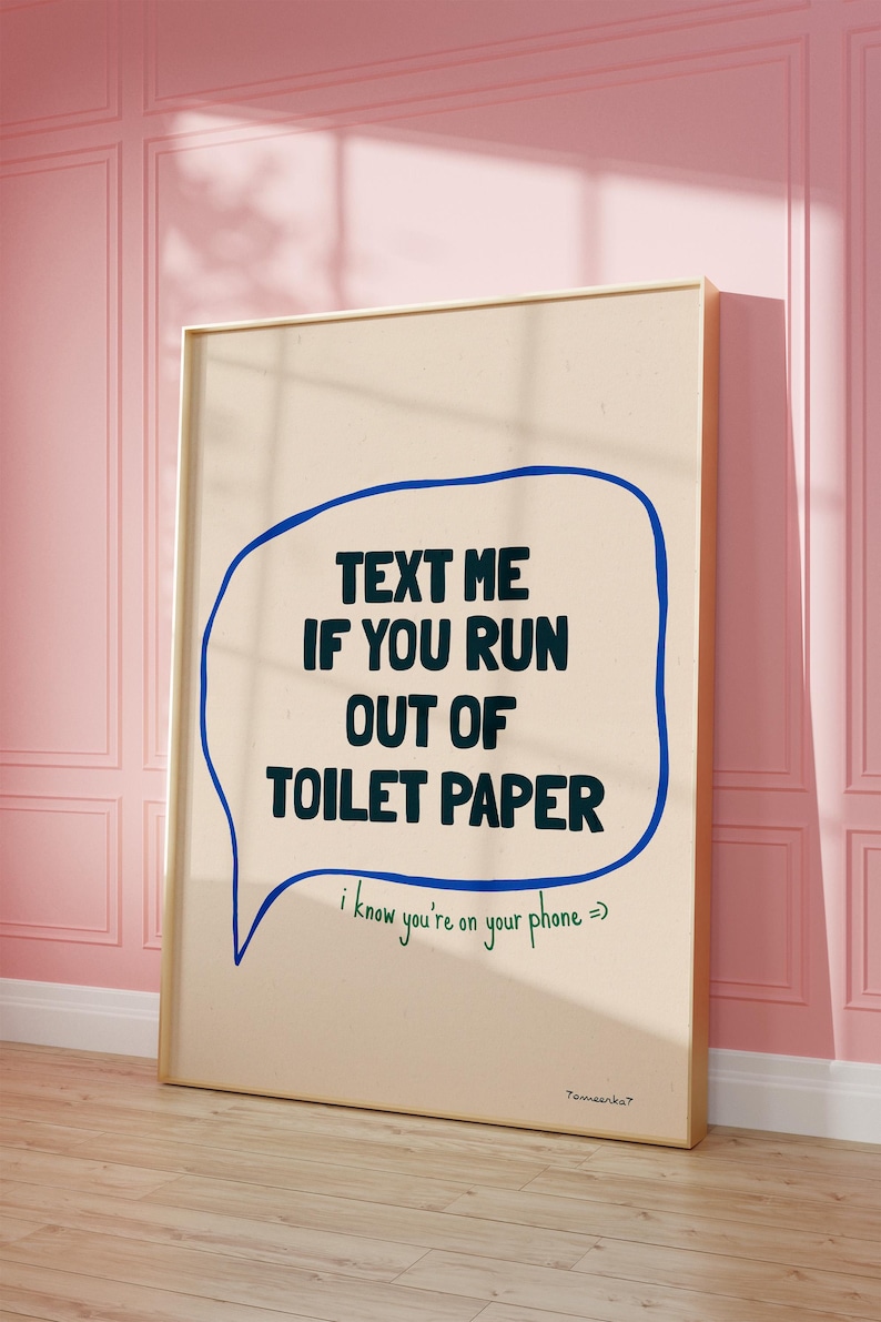 May include: A beige poster with a blue speech bubble that says "TEXT ME IF YOU RUN OUT OF TOILET PAPER" in black text. Below the speech bubble, it says "I know you're on your phone =)" in green text. The poster is framed in gold and is hanging on a pink wall.