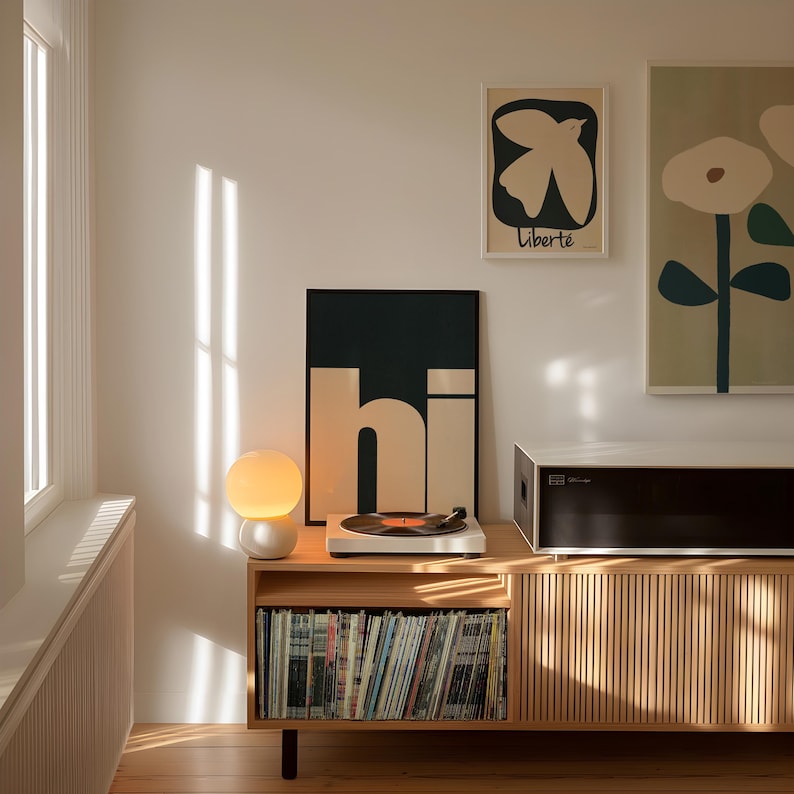 Op de afbeelding: Een houten draaitafelstandaard met een draaitafel, een lamp en een platencollectie. De standaard heeft een lichte houtkleur en een deur met lamellen. Er hangen twee posters aan de muur achter de standaard, een met een vogel en de tekst "Libert&eacute;" en de andere met een gestileerde bloem.