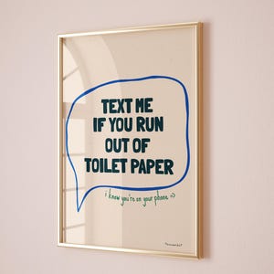May include: A framed print with a blue speech bubble that says "TEXT ME IF YOU RUN OUT OF TOILET PAPER" in black text. The speech bubble has a green handwritten note that says "i know you're on your phone =>". The print has a gold frame and is hanging on a light pink wall.