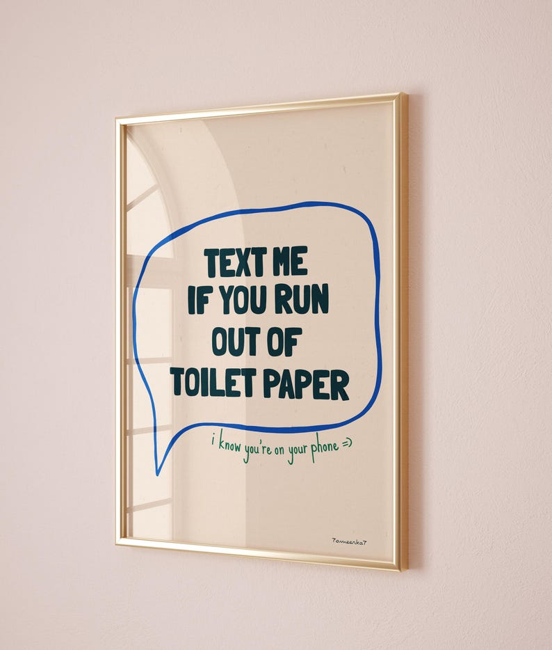 May include: A framed print with a blue speech bubble that says "TEXT ME IF YOU RUN OUT OF TOILET PAPER" in black text. The speech bubble has a green handwritten note that says "i know you're on your phone =>". The print has a gold frame and is hanging on a light pink wall.