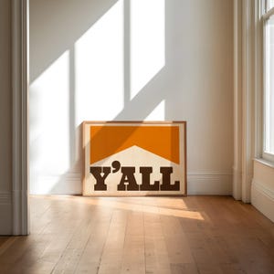 May include: A framed print with a brown and orange color scheme. The print features the text "Y'ALL" in bold, brown letters.