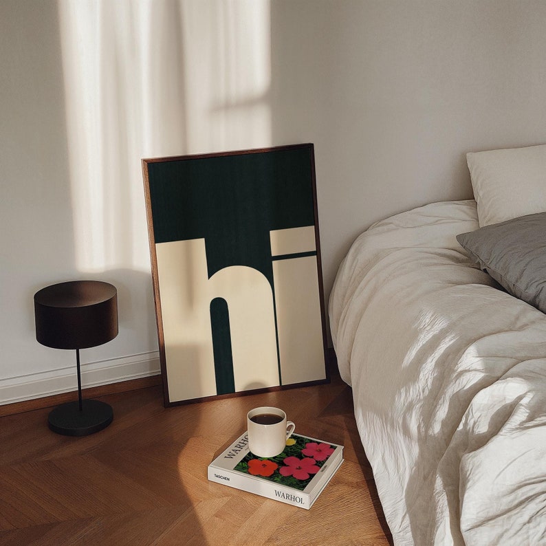 Op de afbeelding: Een ingelijst drukwerk met het woord "hi" in een vet, wit lettertype op een donkergroene achtergrond. De print ligt op een houten oppervlak naast een witte koffiemok en een boek met de titel "Warhol".