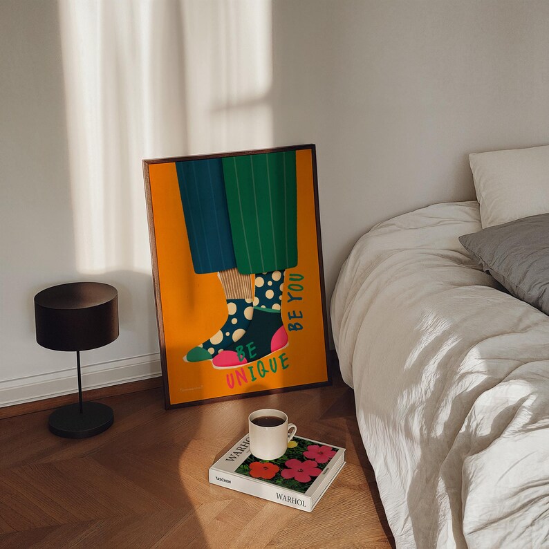 May include: A colorful illustration of legs wearing polka dot socks and the words "Be Unique Be You" on a yellow background. The illustration is framed and hanging on a wall next to a bed with white bedding.