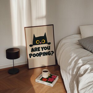 May include: A black cat with yellow eyes peeks over the top of a beige poster with the text "Are you pooping?" The poster is framed in a dark wood frame and is hanging on a white wall. A white bed with a white blanket and a gray pillow is in the background.