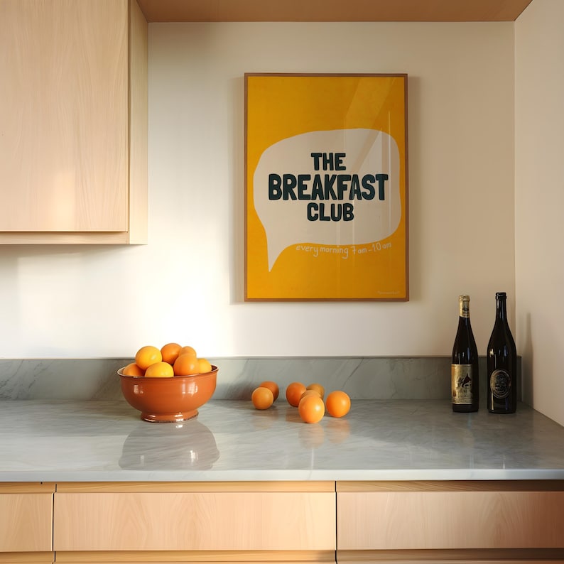 May include: A yellow poster with a white speech bubble that says "The Breakfast Club" in black text. The poster is hanging on a wall in a kitchen with a marble worktop and wooden cabinets. There are oranges in a bowl and two bottles of wine on the counter.