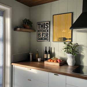Op de afbeelding: Een keuken met witte kasten en een houten aanrecht. Er hangen twee zwart-wit posters aan de muur, een met de tekst "This kitchen is for dancing" en de andere met een geel-zwart gestreept ontwerp. Er staan ook wijnflessen, een vaas met groene planten en een kom met fruit op het aanrecht.