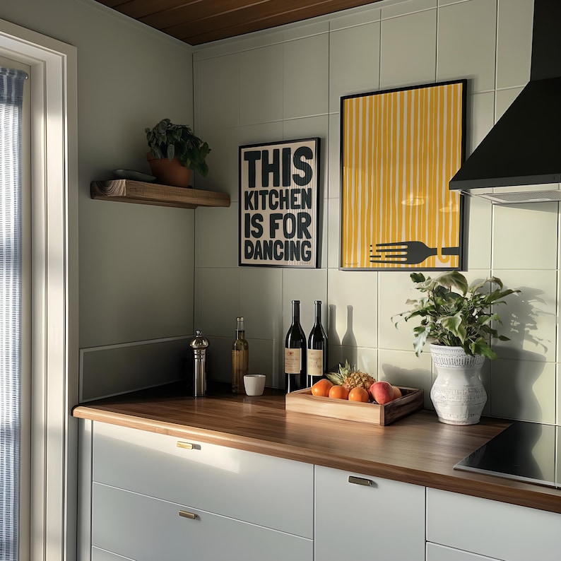 Op de afbeelding: Een keuken met witte kasten en een houten aanrecht. Er hangen twee zwart-wit posters aan de muur, een met de tekst "This kitchen is for dancing" en de andere met een geel-zwart gestreept ontwerp. Er staan ook wijnflessen, een vaas met groene planten en een kom met fruit op het aanrecht.