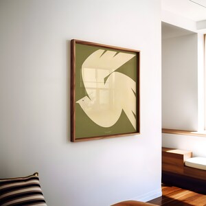Puede incluir: Un cuadro enmarcado con un dise&ntilde;o de paloma blanca estilizada sobre un fondo verde oliva. La obra de arte est&aacute; encerrada en un marco de madera marr&oacute;n oscuro y montada en una pared blanca. El dise&ntilde;o de la paloma es simple y moderno.
