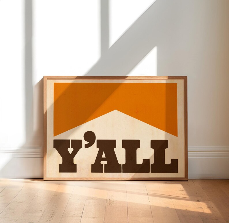 May include: A framed print with a brown wooden frame. The print features a large orange rectangle with a white triangle inside. The text "Y'ALL" is printed in brown letters below the rectangle.