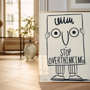 May include: A black and white illustration of a cartoon character holding a sign that says "Stop Overthinking". The character has a simple face with two large eyes and a wide smile. The sign is framed in a gold frame.