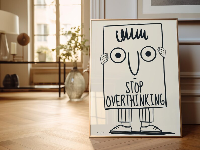 May include: A black and white illustration of a cartoon character holding a sign that says "Stop Overthinking". The character has a simple face with two large eyes and a wide smile. The sign is framed in a gold frame.