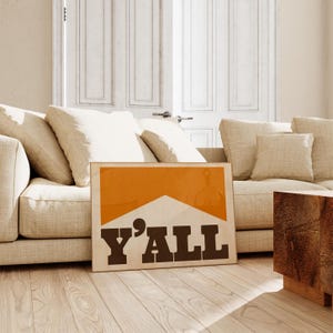 May include: A framed print with an orange background and a white shape resembling a cigarette. The text "Y'ALL" is printed in black letters.