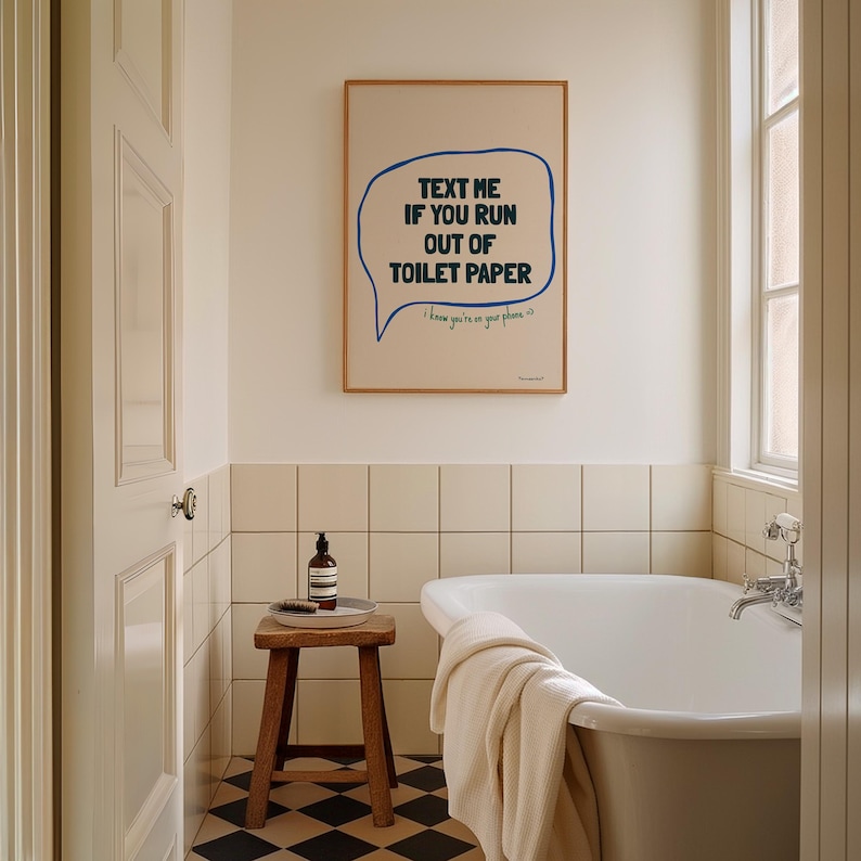 May include: A white bathroom with a black and white checkered floor. A white bathtub with a white towel on the edge. A small wooden stool with a bottle of hand soap on it. A framed print on the wall with the text "Text me if you run out of toilet paper. I know you're on your phone."