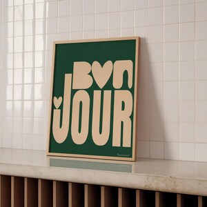 May include: A green framed print with the text "Bon Jour" in a light peach color with hearts in the letters. The print is on a white tiled surface.