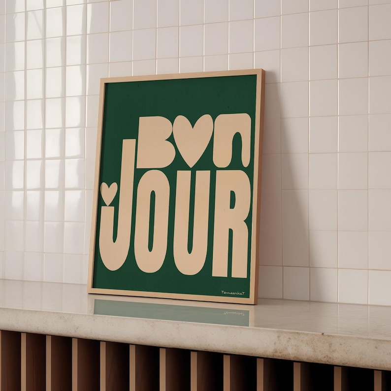 May include: A green framed print with the text "Bon Jour" in a light peach color with hearts in the letters. The print is on a white tiled surface.