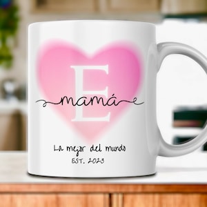 Può includere: Una tazza in ceramica bianca con un design a forma di cuore rosa. Il cuore contiene una grande lettera "E" al centro e la parola "mamá" scritta in corsivo sopra. Il testo "La mejor del mundo EST. 2023" è stampato sotto il cuore.