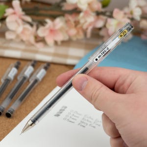 May include: A clear Hi-Tec-C pen with black ink, held over a white sheet of paper. The pen has a 0.25 mm tip. The text "HI-TEC-C" is printed on the side. Several other pens are visible in the background.