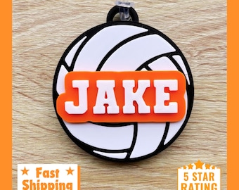May include: White volleyball-shaped luggage tag with a black outline and an orange stripe across the middle. The name "JAKE" is printed in white letters on the orange stripe.