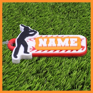 May include: A baseball-themed keyring with a red, white, and orange design. The keyring features a silhouette of a batter and the word "NAME" in white block letters. The background is green.