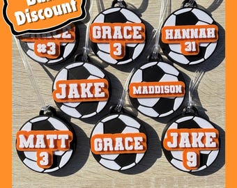 Könnte beinhalten: Acht schwarze und weiße Gepäckanhänger in Form von Fußbällen mit orangefarbenen personalisierten Namen und Nummern. Die Namen sind "Grace", "Hannah", "Jake", "Maddison", "Matt", "Grace" und "Jake". Die Nummern sind "3", "31", "3", "7", "9".